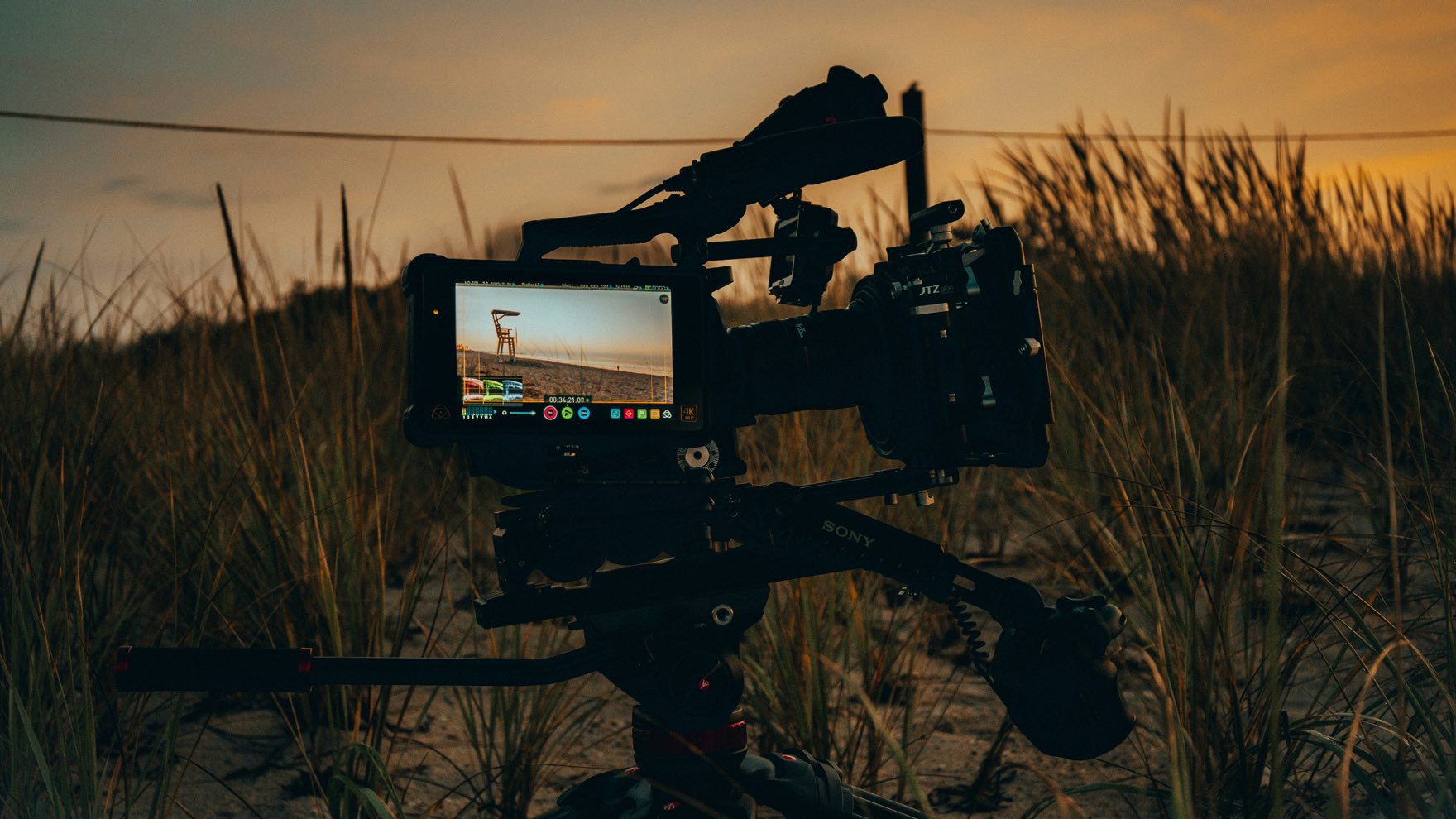A Sony cinema camera rig with an on-board monitor on a tripod in beach dune grass at sunset, framing a lifeguard chair on the sand.